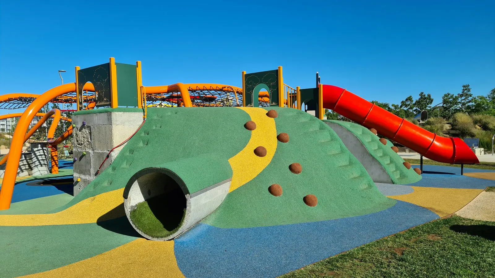 Playground with a green rubber-surfaced climbing hill, a red tube slide and orange climbing arches under a clear blue sky